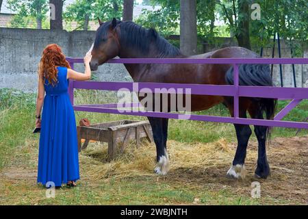 Rothaarige Frau in blauem Kleid streichelt ein Pferd in einem Korral Stockfoto