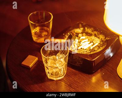 Glas Whiskey mit rauchender Zigarre und Eiswürfeln auf Holztisch Stockfoto