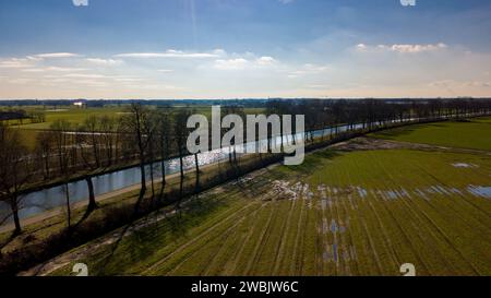 Die Fotografie verkörpert eine ruhige ländliche Landschaft, aufgenommen aus der Luftperspektive. Es verfügt über einen glitzernden Fluss, der die Sonnenstrahlen einfängt, umgeben von einer Reihe von Bäumen, die lange Schatten über das Gelände werfen. Das angrenzende Ackerland ist mit verschiedenen Grüntönen durchzogen, was auf frühe Vegetation hinweist, wobei einige Bereiche stehendes Wasser zeigen, das den Himmel reflektiert, möglicherweise durch jüngste Regenfälle. Der ferne Horizont ist von den Silhouetten von Gebäuden gesäumt, die auf eine nahe gelegene Gemeinde hinweisen. Die Sonnenstrahlen, die durch die Wolken dringen, verleihen der ansonsten ruhigen Landschaft ein dynamisches Element. Stockfoto