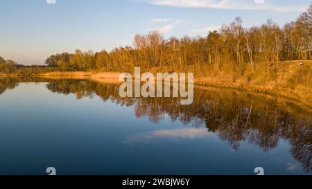 Dieses ruhige Bild fängt das ruhige Wasser eines Flusses während der goldenen Stunde ein, während das warme Licht der untergehenden Sonne die Bäume und Gräser entlang der Ufer beleuchtet. Die Bäume, die von ihren Blättern befreit sind, zeigen komplizierte Astmuster, die sich perfekt in der gläsernen Oberfläche des Flusses spiegeln und ein symmetrisches Naturkunstwerk schaffen. Die sanften Goldtöne und der klare blaue Himmel verleihen der Szene ein Gefühl von ruhiger Wärme, was auf einen späten Herbst oder frühen Wintertag hinweist. Dieses Foto lädt zum Nachdenken und zu einer tiefen Wertschätzung für den friedlichen Fluss der Natur ein. Goldene Stunde Reflexion über einen Tra Stockfoto