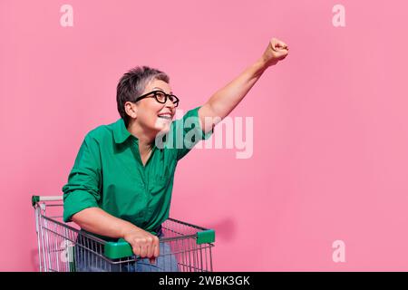 Porträt der optimistischen Person tragen Jeanshosen, die auf Trolley reiten, Raisng Faust-Blick leerer Raum isoliert auf rosa Hintergrund Stockfoto