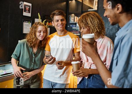 Trendiger Mann mit Smartphone in der Nähe fröhlicher multikultureller Freunde an der Rezeption in einem modernen Hostel Stockfoto