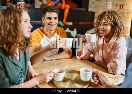 Fröhliche multikulturelle Freunde halten Kaffeetassen und unterhalten sich im Hostel-Café, reisen und Freundschaft Stockfoto