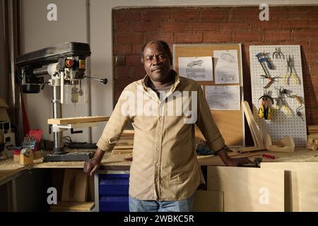 Afroamerikanischer hochrangiger Handwerker, der in der sonnendurchfluteten Werkstatt neben den Maschinen steht Stockfoto