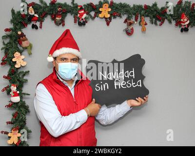 Lateinischer Mann mit Schutzmaske, Weihnachtsmütze und Tafel mit der Botschaft "Weihnachten ohne Party" in spanischer Sprache. Weihnachtsdekoration, neu normal Stockfoto