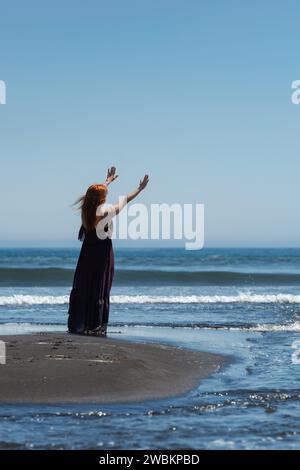 Frau in lila Kleid sieht so glücklich und sorglos aus, wie sie am Sandstrand steht, mit hoch erhobenen Armen zum wunderschönen blauen Himmel und den Meereswellen Stockfoto