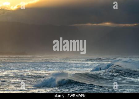 Ein einsamer Surfer reitet in Ho'okipa auf den gewaltigen Wellen, während der Sonnenuntergang auf Maui den Himmel mit Dramen übermalt. Stockfoto