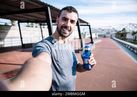 Runner man trinkt Wasser und macht ein Selfie nach dem Training draußen. Gutaussehender Sportmann, der eine Flasche hält, ein Foto macht und in die Kamera schaut. Stockfoto