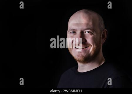 Porträt eines lächelnden erwachsenen Glatzenmannes auf schwarzem Hintergrund. Kopierbereich für den Text. Nahaufnahme Porträt eines glücklichen weißen Mannes in leiser Sprache. Stockfoto