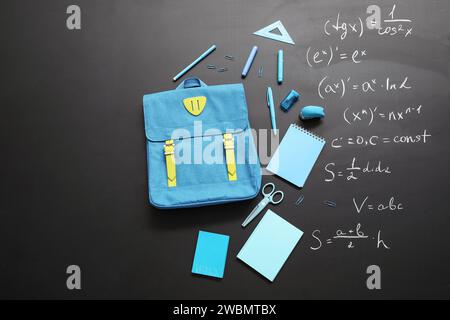 Blauer Schulrucksack mit verschiedenen Schreibwaren auf schwarzer Tafel, mit Formeln bemalt Stockfoto