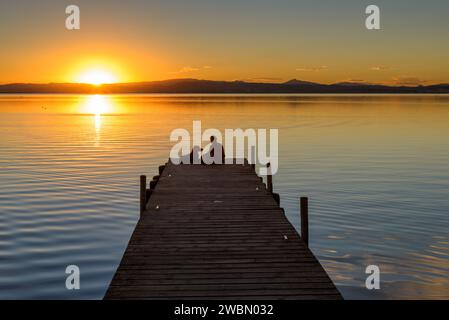 Ein Mädchen und ein Hund sitzen auf dem Steg des Albufera-Sees und beobachten einen farbenfrohen und wunderschönen Sonnenuntergang in Valencia, Spanien. Stockfoto