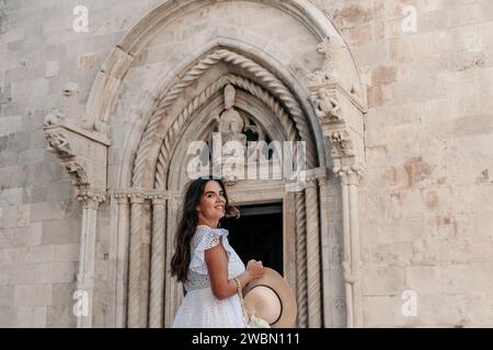 Eine elegante junge Frau in einem fließenden weißen Kleid posiert anmutig vor einer majestätischen, historischen Kirche Stockfoto