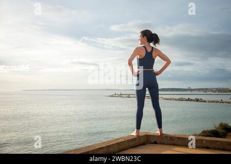 Rückansicht einer sportlichen Frau mit Blick auf das Meer mit Händen auf den Hüften, entspannend nach dem Training. Stockfoto