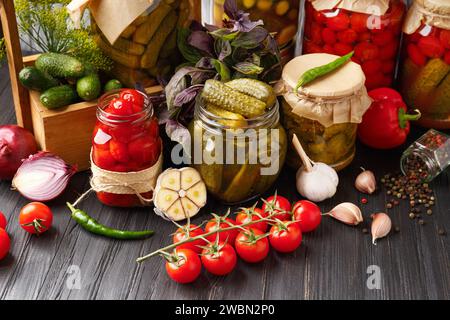 Konservierte Kirschtomaten und Gurken in Gläsern, frisches Gemüse, Gewürze und Kräuter für Marinade auf hölzernem Hintergrund. Eingelegtes Gemüse. Stockfoto