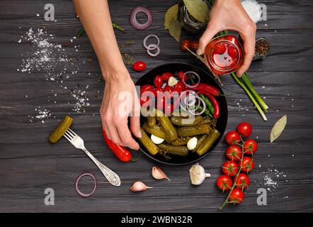 Eingelegte Kirschtomaten, Gurken auf schwarzem Teller und Frauenhände legen Gemüse auf dunklem Holzhintergrund von oben hin. Stockfoto