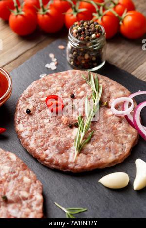 Rohe frisch gemahlene Rindfleisch-Burger-Patties, Rosmarin, Gemüse und Gewürze auf einem schwarzen Steinteller auf hölzernem Hintergrund, Nahaufnahme. Stockfoto