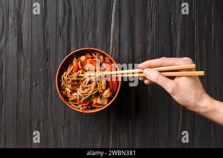 Teller mit Buchweizennudeln mit Gemüse, Pilzen, Hühnerfleisch auf dunklem Hintergrund. Chinesische Stöcke nehmen japanische Soba aus einer Tonschüssel Stockfoto