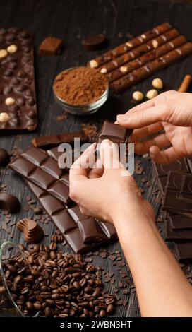 Schokolade verschiedener Art, Kakaopulver, Kaffeebohnen, Haselnüsse auf dunklem Holzhintergrund. Mädchenhände brechen Schokolade, Konzept der Konditorin Stockfoto