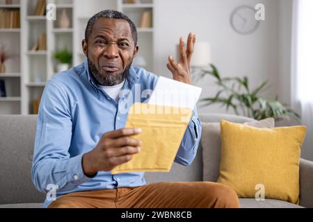 Porträt eines besorgten afroamerikanischen Mannes, der zu Hause auf der Couch sitzt und einen Umschlag mit einem Brief in der Hand hält und frustriert seine Hände ausbreitet und in die Kamera blickt. Stockfoto