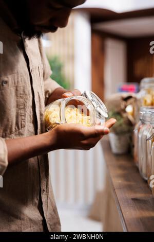 Nahaufnahme eines schwarzen Mannes, der frisches, lokal angebautes Getreide und Nudeln untersucht. Selektiver Fokus eines afroamerikanischen männlichen Kunden, der ein Glas mit Spaghetti hält und das hausgemachte Produkt bewundert. Stockfoto