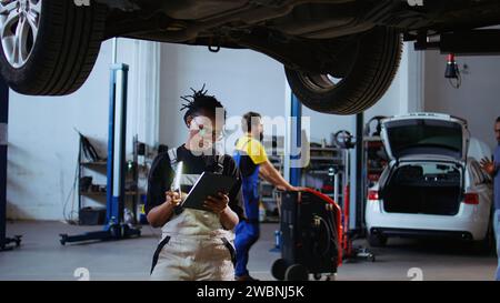 Akribischer Techniker unter dem hängenden Fahrzeug in der Werkstatt, der während der Wartung mit einem Arbeitsscheinwerfer auf Schäden überprüft. Werkstattarbeiter mit professionellem Werkzeug, um das Fahrzeug auf einer Deckenaufhängung zu untersuchen Stockfoto