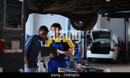 Geschulter Mechaniker und Kunde, der unter dem Auto in der Garage mit Laptop steht, um neue Teile zu bestellen, nachdem Mängel entdeckt wurden. Mitarbeiter, der afroamerikanischen Kunden hilft, indem er online nach Komponenten sucht Stockfoto