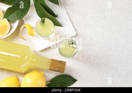 Leckerer Limoncello-Likör, Zitronen und grüne Blätter auf einem hellen, strukturierten Tisch, flach gelegen. Leerzeichen für Text Stockfoto