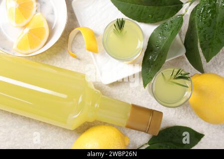 Leckerer Limoncello-Likör, Zitronen und grüne Blätter auf einem hellen, strukturierten Tisch, flach gelegen Stockfoto