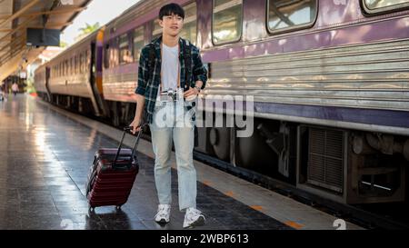 Ein glücklicher, gutaussehender junger asiatischer Mann mit Rucksack und Gepäck, der den Zug an einem Bahnhof nimmt, auf einem Bahnsteig läuft, irgendwo an einem reist Stockfoto