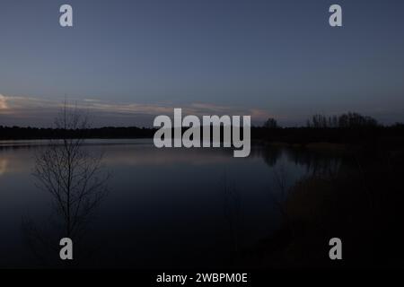 In diesem Bild ist die Ruhe des frühen Abends spürbar, wenn das letzte Licht des Tages vom Himmel vergeht, gespiegelt von der stillen Oberfläche eines ruhigen Sees. Die Silhouetten von jungen Bäumen und Sträuchern ohne Blätter lassen die Kühle des Spätherbst oder frühen Winters vermuten. Es gibt einen sanften Verlauf am Himmel vom Dämmerungsblau bis zur restlichen Wärme des Sonnenuntergangs, der einen sanften nächtlichen Tag bietet. Die Stille des Wassers deutet auf einen Moment der Pause hin, eine natürliche Einladung, über den vergangen Tag nachzudenken, wenn sich die Nacht nähert. Abendliche Ruhe auf einem stillen See. Hochwertige Fotos Stockfoto