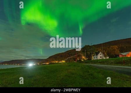 Wunderschöne grüne Nordlichter über einem Fjord und einem Haus auf der Insel Kvaloya in der Nähe von Tromsø. Tanzende Polarlichter über einem Berg, aurora borealis Stockfoto