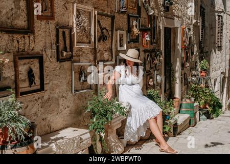 Eine elegante Frau, die ein makelloses weißes Kleid und einen Hut trägt und sich in einer malerischen Gasse entspannt Stockfoto