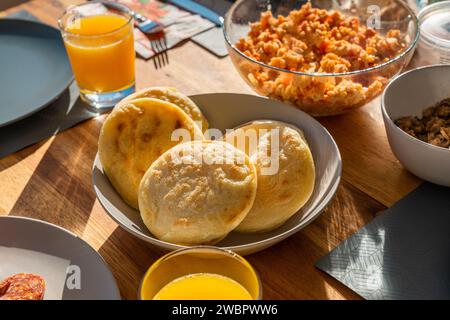 Arepas in der Mitte des Frühstückstisches neben Rührei, Fleisch und Orangensaft in direkter Sonneneinstrahlung. Stockfoto