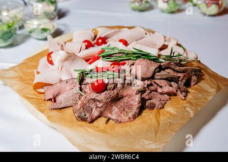 Eine Gourmet-Fleischplatte mit saftig geschnittenem Roastbeef und truthahn, garniert mit Kirschtomaten und frischem Rosmarin auf einem rustikalen, papierbeschichteten Holz Stockfoto