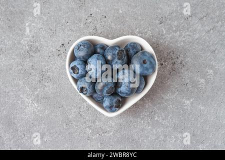 Frische Heidelbeeren in einer kleinen herzförmigen Schüssel auf grauem Betonhintergrund. Bio-Beeren, gesundes Essen, wilde Beeren. Draufsicht, flach Stockfoto