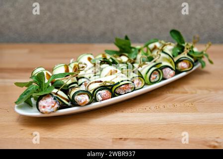 Delikate Zucchini-Roll-ups gefüllt mit Kräuterkäse und geräuchertem Lachs, garniert mit frischem Gemüse, präsentiert auf einer weißen Platte. Stockfoto