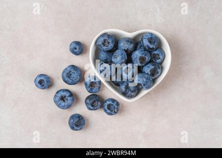 Frische Heidelbeeren in einer kleinen herzförmigen Schüssel auf beigefarbenem Betonhintergrund. Bio-Beeren, gesundes Essen, wilde Beeren. Draufsicht, flach Stockfoto