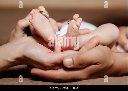 Mom und Dad halten die Beine ihres Kindes, goldene Eheringe an den Zehen. Stockfoto