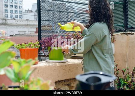 Junge Frau mit Lockenwicklern, die im Garten auf der Terrasse ihres Hauses steht, Wasser auf Pflanzen mit Plastikbehälter legt, Wartungsarbeiten macht und Auto nimmt Stockfoto