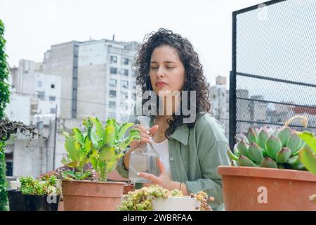 Junge lateinfrau in Lockenwicklern, die bei der Arbeit steht und Pflanzen auf der Terrasse pflegt, Wasserspritze benutzt, um sie zu hydrieren, Hausarbeitskonzept, Kopierraum. Stockfoto