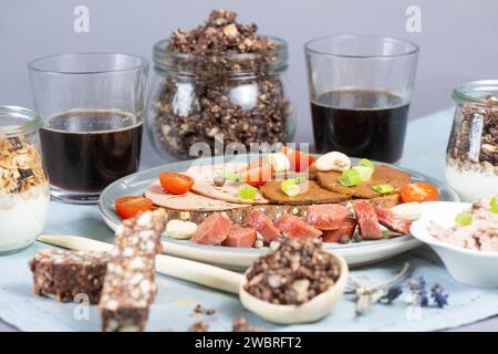 Dresden, Deutschland. Januar 2024. Eine Brotscheibe mit verschiedenen veganen Würstchen, eine vegane Knoblauchwurst und eine Schüssel mit einer veganen Leberwurstalternative auf Erbsenbasis, Müsli mit pflanzlicher Joghurtalternative, Cashew Dattelbar und Kaffee auf einem Tisch. Quelle: Sebastian Kahnert/dpa/Alamy Live News Stockfoto