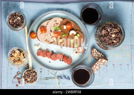 Dresden, Deutschland. Januar 2024. Eine Brotscheibe mit verschiedenen veganen Würstchen, eine vegane Knoblauchwurst und eine Schüssel mit einer veganen Leberwurstalternative auf Erbsenbasis, Müsli mit pflanzlicher Joghurtalternative, Cashew Dattelbar und Kaffee auf einem Tisch. Quelle: Sebastian Kahnert/dpa/Alamy Live News Stockfoto