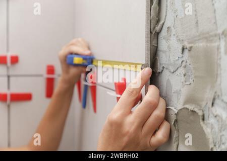 Frau fliesst Badezimmerwände mit Bandmaß zum Messen der Länge. Der Prozess der schrittweisen Installation von Fliesen im Badezimmer. Heimwerkerimprovisieren Stockfoto