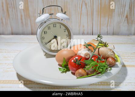 Intervallfasten als gesundes Diät-Konzept zum Abnehmen, weiße Tafel gefüllt zu einem Drittel mit Nahrung und Wecker auf einer leichten rustikalen Holzbaa Stockfoto