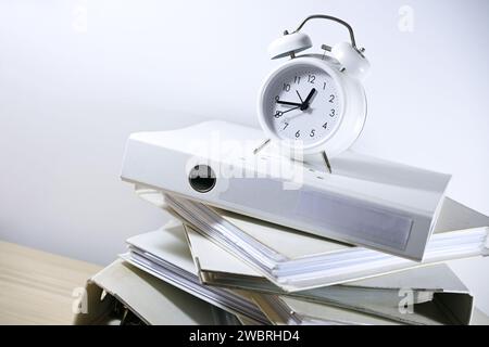 Schräg gestapelte Ringordner und ein weißer Wecker oben, der an die Frist erinnert, Geschäftskonzept von Bürokratie, Verwaltung, Arbeitsüberlastung Stockfoto