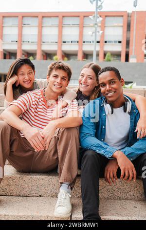 Vertikales Porträt einer Gruppe von echten multirassischen Schülern, die lächelnd und in die Kamera schauen, die auf den Stufen einer Universitätscampustreppe sitzt. Internationale Teenager-Freunde lachen. Hochwertige Fotos Stockfoto