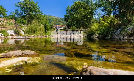 Klarer Fluss mit Steinen, umgeben von Vegetation, und Häuser einer kleinen ländlichen Stadt in der Ferne. Carmona, Kantabrien, Spanien. Stockfoto