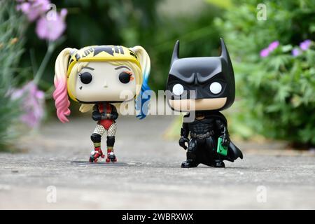 Funko Pop Actionfiguren der DC-Comic-Superhelden Harley Quinn und Batman stehen auf Asphaltstraße im Spring Park, grünem Gras und rosa Blumen. Stockfoto