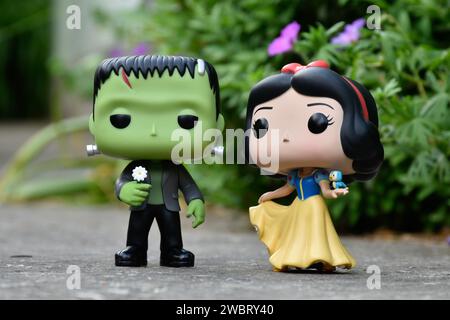 Funko Pop Actionfiguren von Frankensteins Monster mit Gänseblümchen und Disney Prinzessin Schneewittchen mit Vogel. Asphaltstraße, rosa Blumen, grünes Gras. Stockfoto