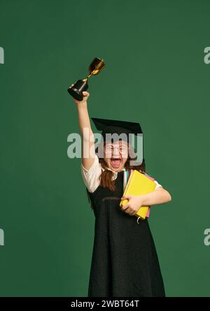 Glückliches Mädchen mit Down-Syndrom, das in der Abschlussmütze, Büchern und Trophäe vor grünem Studiohintergrund steht Stockfoto